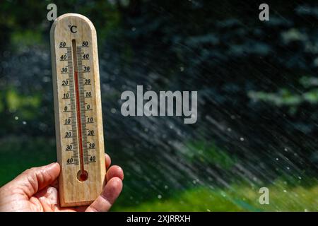 Un uomo tiene in mano un termometro di legno in giardino che indica una temperatura molto elevata - protezione contro il calore Foto Stock