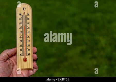 Un uomo tiene in mano un termometro di legno in giardino che indica una temperatura molto elevata - protezione contro il calore Foto Stock