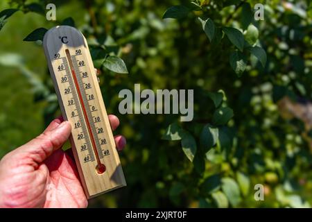 Un uomo tiene in mano un termometro di legno in giardino che indica una temperatura molto elevata - protezione contro il calore Foto Stock