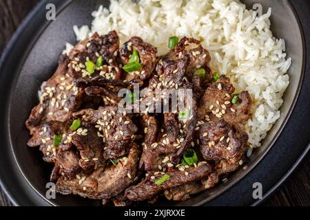 Bulgogi, manzo BBQ coreano, manzo fritto a fette sottili cosparso di semi di sesamo e cipolla di primavera servito con riso a grani lunghi in una ciotola nera, primo piano Foto Stock