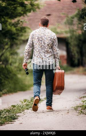 Un uomo cammina lungo un sentiero di ghiaia in campagna, portando con sé una valigia marrone e occhiali da sole. Indossa una camicia e un jeans a motivi geometrici. Foto Stock