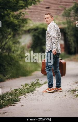 Un uomo con una camicia floreale e un jeans si allontana su una strada di campagna, portando una valigia marrone. Foto Stock