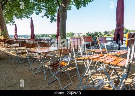 Una birreria all'aperto vuota con tavoli e sedie all'ombra degli alberi in una giornata di sole, con vista sul fiume Reno a Neuwied, in Germania. Foto Stock