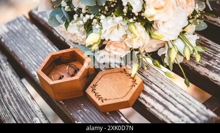 Due fedi nuziali riposano in una scatola di legno accanto a un mazzo di fiori bianchi su una panchina di legno intemprata. Foto Stock