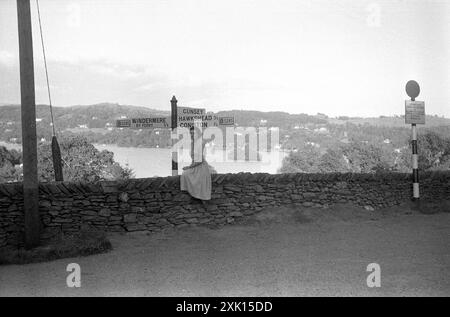 Lontano Sawrey, Cumbria. 1957 – Una donna in posa per una fotografia sedeva su un muro di pietra che si affacciava sul lago Windermere. Si trova sulla strada B5285 a far Sawrey in Cumbria, Inghilterra, vicino al traghetto Windermere. Questo traghetto trasporta passeggeri e veicoli attraverso il lago Windermere tra far Sawrey e Bowness su Windermere. Dietro c'è un cartello stradale che fornisce indicazioni per Windermere, Cunsey, Hawkshead e Consiton. Foto Stock