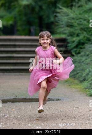 Una bambina carina in un bellissimo vestito rosa con capelli lunghi corre rapidamente. Giochi all'aperto. Sfondo sfocato. Foto Stock