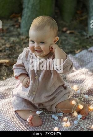 Ritratto di un affascinante bambino di un anno in un abito beige, seduto vicino a un grande albero. Il bambino tiene le lanterne tra le mani e le esamina attentamente Foto Stock