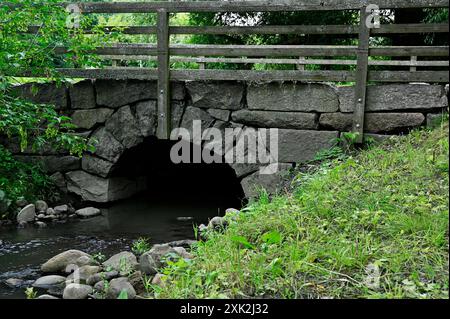 Un ponte rustico in pietra con ringhiera in legno si estende su un piccolo torrente circondato da vegetazione lussureggiante e rocce. L'arco del ponte e l'ambiente naturale Foto Stock