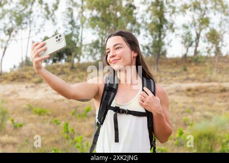 Una giovane escursionista sorridente scatta un selfie con il suo smartphone in mezzo a un paesaggio naturale, mostrando la sua avventura all'aria aperta e il suo legame con natu Foto Stock