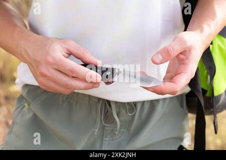 Immagine ravvicinata delle mani di un escursionista mentre maneggia con cura un coltello nero pieghevole durante un'escursione in campagna, mettendo in evidenza il surv all'aperto Foto Stock