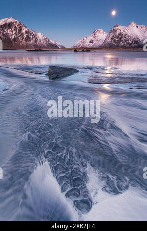 Un tranquillo paesaggio delle Lofoten sotto la luna, caratterizzato da maree ghiacciate e montagne innevate che si riflettono in acque calme Foto Stock