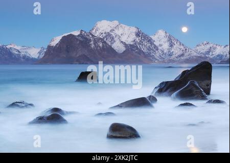 Una tranquilla scena al chiaro di luna cattura la bellezza serena di Lofoten, con montagne innevate e superfici d'acqua lisce, accentuate da una luna scintillante Foto Stock