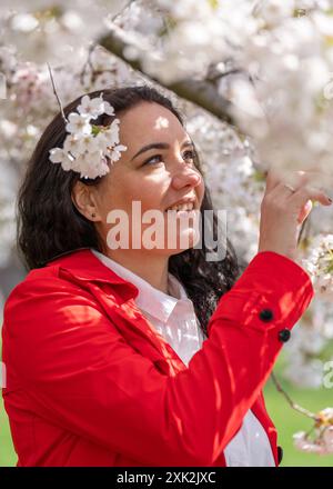 immagine romantica di una donna elegante con un cappotto rosso, in una camicetta bianca. Umore positivo. Una ragazza carina tiene delicatamente un ramo di sakura bianca e guarda il Foto Stock