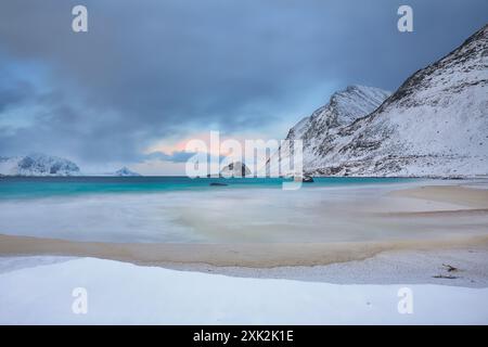 Un sereno crepuscolo invernale a Haukland Beach a Lofoten, Norvegia, con acque turchesi lisce, una spiaggia sabbiosa innevata e aspre montagne sotto pa Foto Stock