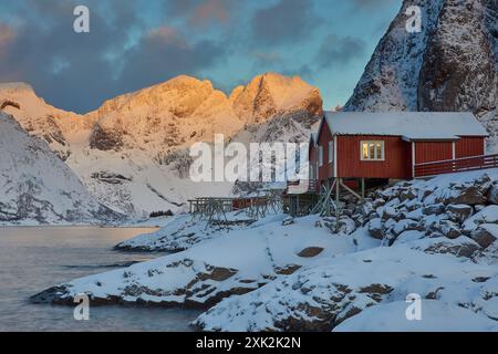 Un'incantevole alba invernale illumina le cime innevate e una tradizionale cabina di pesca rossa a Lofoten, Norvegia, che riflette una terra nordica mozzafiato Foto Stock