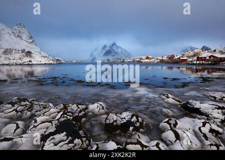 Un tranquillo scenario invernale a Lofoten, Norvegia, caratterizzato da piccole case colorate accanto a una baia tranquilla circondata da montagne innevate sotto un blu moody Foto Stock