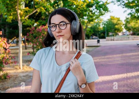 Una giovane donna con occhiali e cuffie ascolta la musica mentre cammina in un parco. Indossa una camicetta bianca casual e porta con sé una borsa a spalla. Foto Stock
