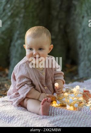 Ritratto di una bambina affascinante in un abito beige seduta vicino a un grande albero, guardando la macchina fotografica. Tiene le lanterne e le mastica con piacere. Foto Stock