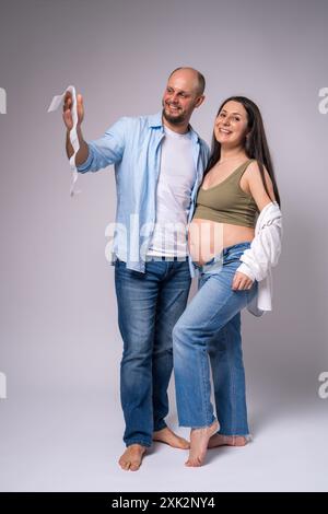 Una donna incinta con una camicia bianca, jeans blu con la pancia esposta e suo marito stanno tenendo in mano una foto del loro neonato non ancora nato. Gravidanza, falena Foto Stock