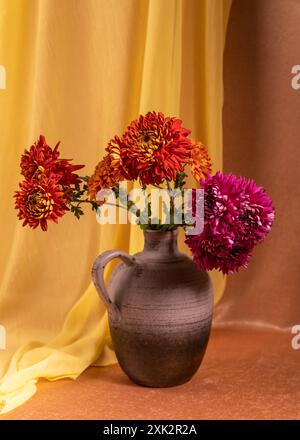 Un bouquet di dahlias rosse e rosa in un vaso su sfondo arancione, accanto ad esso si trova una sciarpa gialla. Natura morta, cartolina Foto Stock