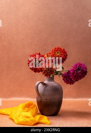 Un bouquet di dahlias rosse e rosa in un vaso su sfondo arancione, accanto ad esso si trova una sciarpa gialla. Natura morta, cartolina Foto Stock