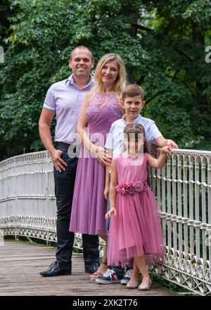 Ritratto di una famiglia felice. Papà e mamma ridendo, figlio adolescente e figlia di prima elementare stanno su un ponte bianco nel parco. Mamma e ragazza sono vestite Foto Stock