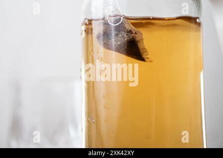 Primo piano di una bottiglia di vetro riempita di liquido color ambra, probabilmente tè o whisky, con una bustina di tè che si insinua all'interno. Lo sfondo è sfocato, emp Foto Stock