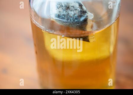 Un primo piano di un vaso di vetro contenente un liquido dorato, probabilmente tè, con una bustina di tè immersa all'interno. Il vaso è posizionato su una superficie di legno, in modo da mostrare Foto Stock