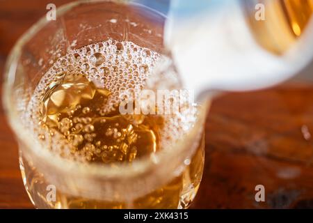 Primo piano di un bicchiere riempito di una bevanda dorata frizzante, versato da una caraffa. La bevanda ha un aspetto frizzante con bollicine che salgono a t Foto Stock