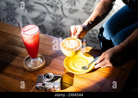 Un'accogliente caffetteria con una persona che tiene una tazza gialla di latte art su un piattino, accanto a un alto bicchiere di bevanda rossa. Una vecchia fotocamera si appoggia sulla W. Foto Stock