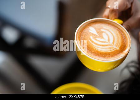 Primo piano di una mano che tiene in mano una tazza di caffè giallo con sopra una latte art. Lo sfondo e' sfocato, presenta un intimo ambiente da caffe' con illuminazione soffusa. Foto Stock