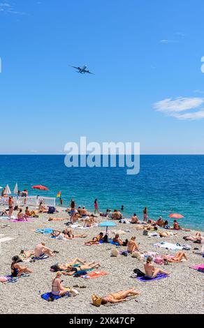 Le persone su una spiaggia di ciottoli lungo il lungomare di Nizza mentre un aereo vola in alto, Nizza, Provence-Alpes-Cote d'Azur, Alpes-Maritimes, Francia Foto Stock