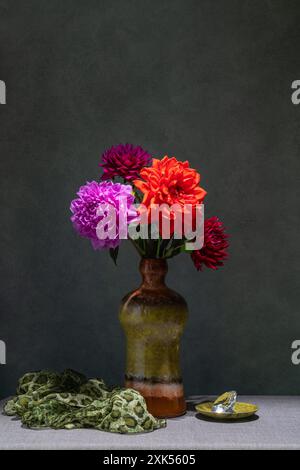 Natura morta con fiori. Dahlias rosa e rosse in vaso scuro, tessuto rosa, piccola tazza rosa, sfondo scuro Foto Stock