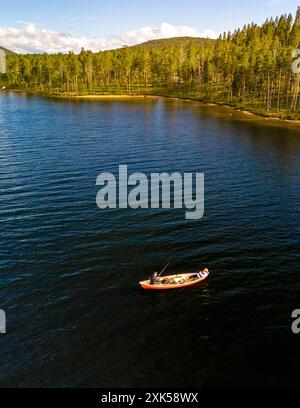 Un pescatore solitario getta la sua linea da una piccola barca nelle acque tranquille di un lago Lapponia finlandese, circondato da una vegetazione lussureggiante e da un cielo blu brillante. Foto Stock