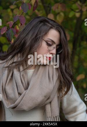 Ritratto di una bella ragazza dai capelli scuri di 20 anni che indossa gli occhiali in un parco autunnale, da vicino. La ragazza ha guardato in basso con attenzione. Femini Foto Stock