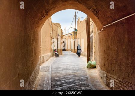 Donna iraniana che indossa un chador nero che cammina lungo le strette strade della storica città di Yazd. Architettura persiana unica della città antica. Foto Stock