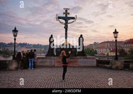 Una donna solitaria fotografa l'alba dal Ponte Carlo di Praga, davanti alla statua di Cristo sulla Croce, un gruppo di amici si accoccolano nelle vicinanze Foto Stock