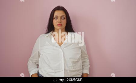 Bellissima giovane donna taglia Plus in piedi su uno sfondo rosa con un'espressione sicura che indossa una camicia bianca e un paio di jeans Foto Stock