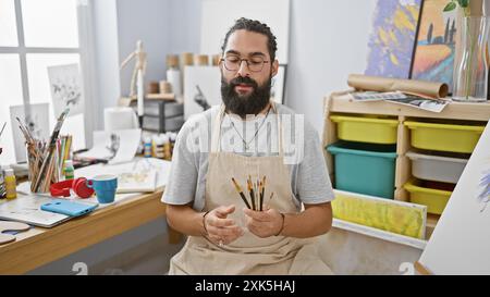 Un uomo barbuto con occhiali che reggono pennelli in un luminoso studio d'arte. Foto Stock