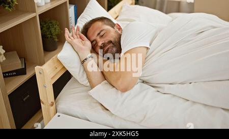 Un uomo maturo e barbuto in abiti bianchi dorme pacificamente in una camera da letto accogliente e ben arredata, evocando comfort e tranquillità. Foto Stock