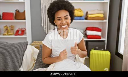 Una allegra donna afroamericana con capelli ricci regala due pollici mentre si siede in una camera da letto moderna. Foto Stock