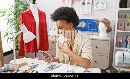 Donna africana sorridente che utilizza lo smartphone in una stanza dal design colorato con manichino e articoli da cucire. Foto Stock