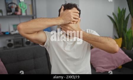 Un uomo in difficoltà copre il suo viso con le mani in un salotto moderno, trasmettendo emozioni di stress o tristezza. Foto Stock
