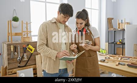 Un uomo e una donna, entrambi falegnami, collaborano in un'officina ben attrezzata facendo riferimento a una nota in un notebook. Foto Stock