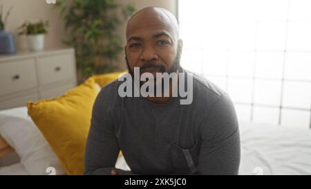 Bel giovane uomo nero con la barba seduto su un letto in un'accogliente camera da letto con piante da interno e cuscini gialli, creando una casa calda e confortevole Foto Stock