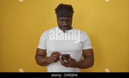 Bell'afroamericano con la barba che indossa una camicia bianca e un cerchietto, che utilizza uno smartphone su uno sfondo giallo isolato. Foto Stock