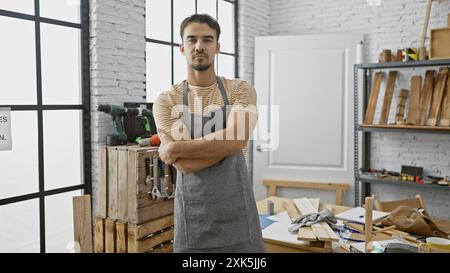 Giovane sicuro di sé con la barba in piedi in un laboratorio di falegnameria ben illuminato, braccia incrociate. Foto Stock