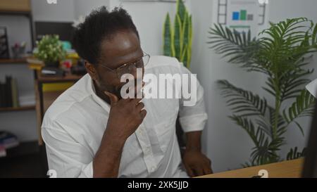 Giovane afro-americano con la barba in un ambiente d'ufficio, meditando guardando il suo computer, con piante e scaffali sullo sfondo. Foto Stock