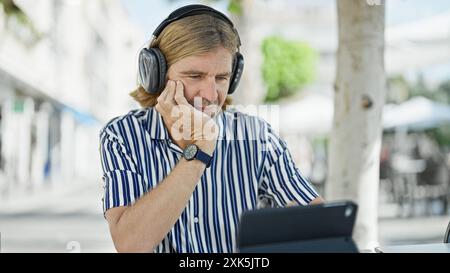 Un uomo pensivo con le cuffie usa un tablet in un bar all'aperto in città. Foto Stock