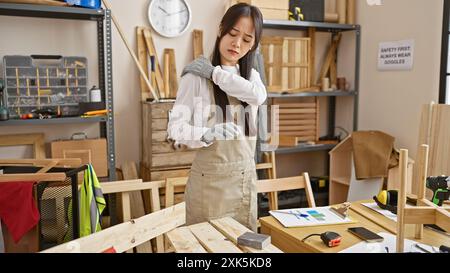 Una donna asiatica soffre di mal di schiena mentre lavora in un laboratorio con legno, attrezzi e attrezzature di sicurezza. Foto Stock